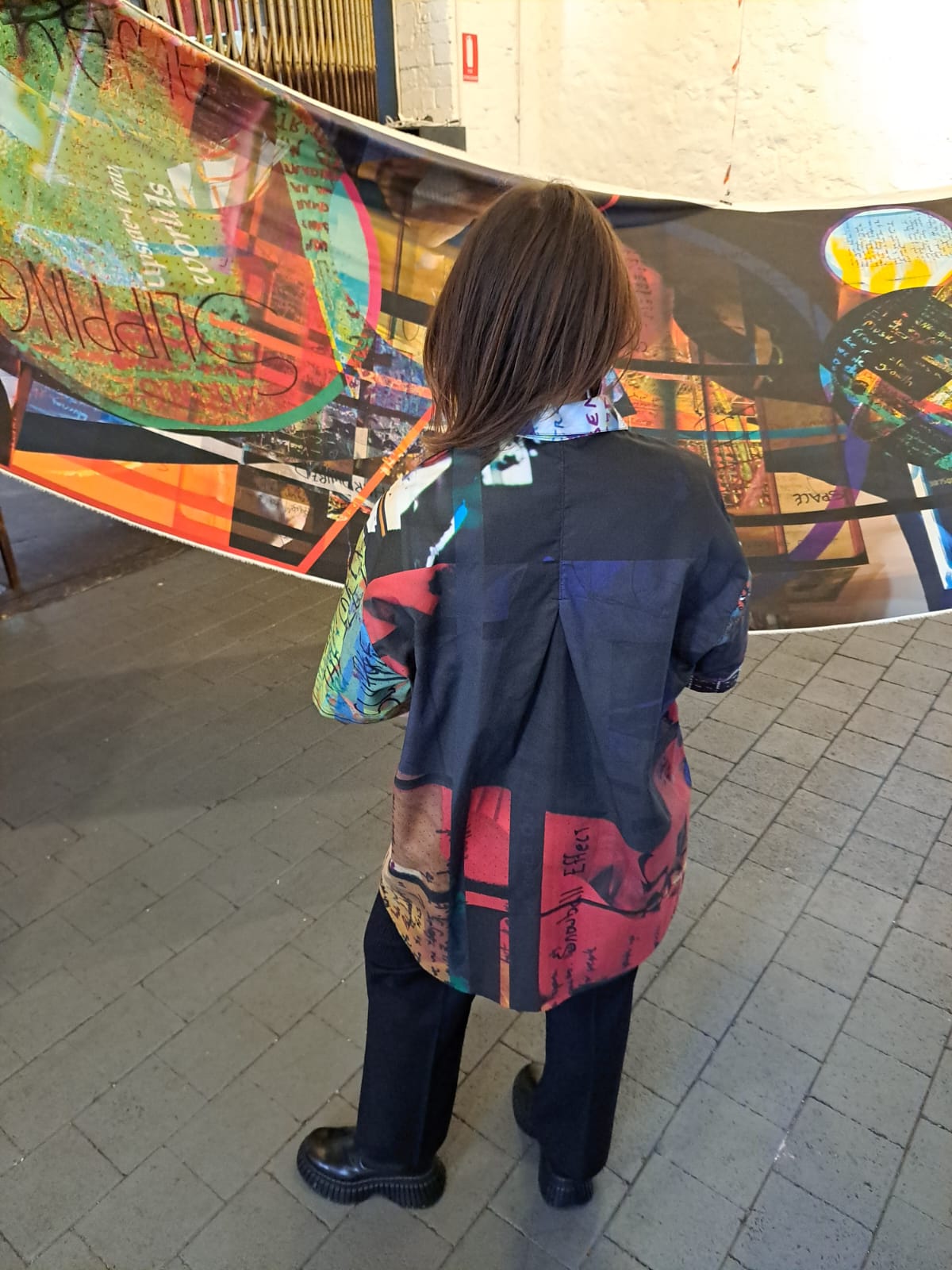 Person wearing a printed short sleeved shirt, facing away from the camera, standing in front of a colorful fabric print indoors