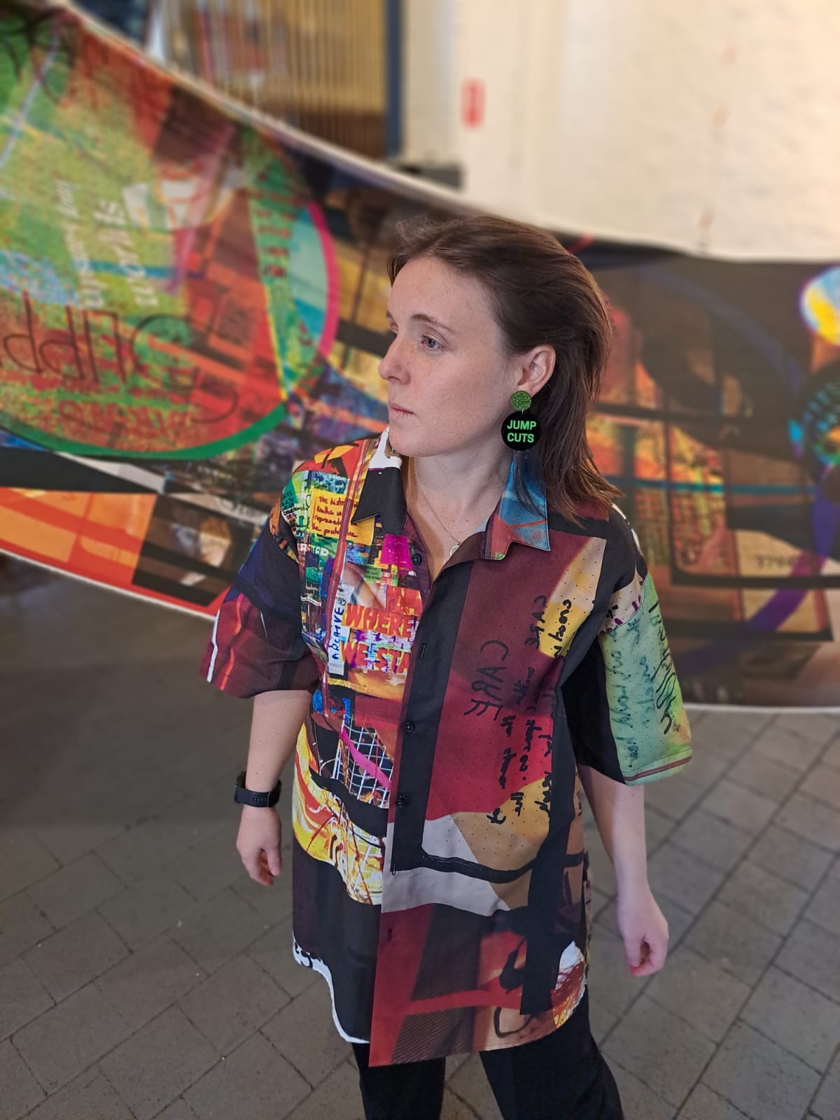 Woman wearing a colorful patterned shirt with abstract designs, standing in front of a large abstract fabric print.