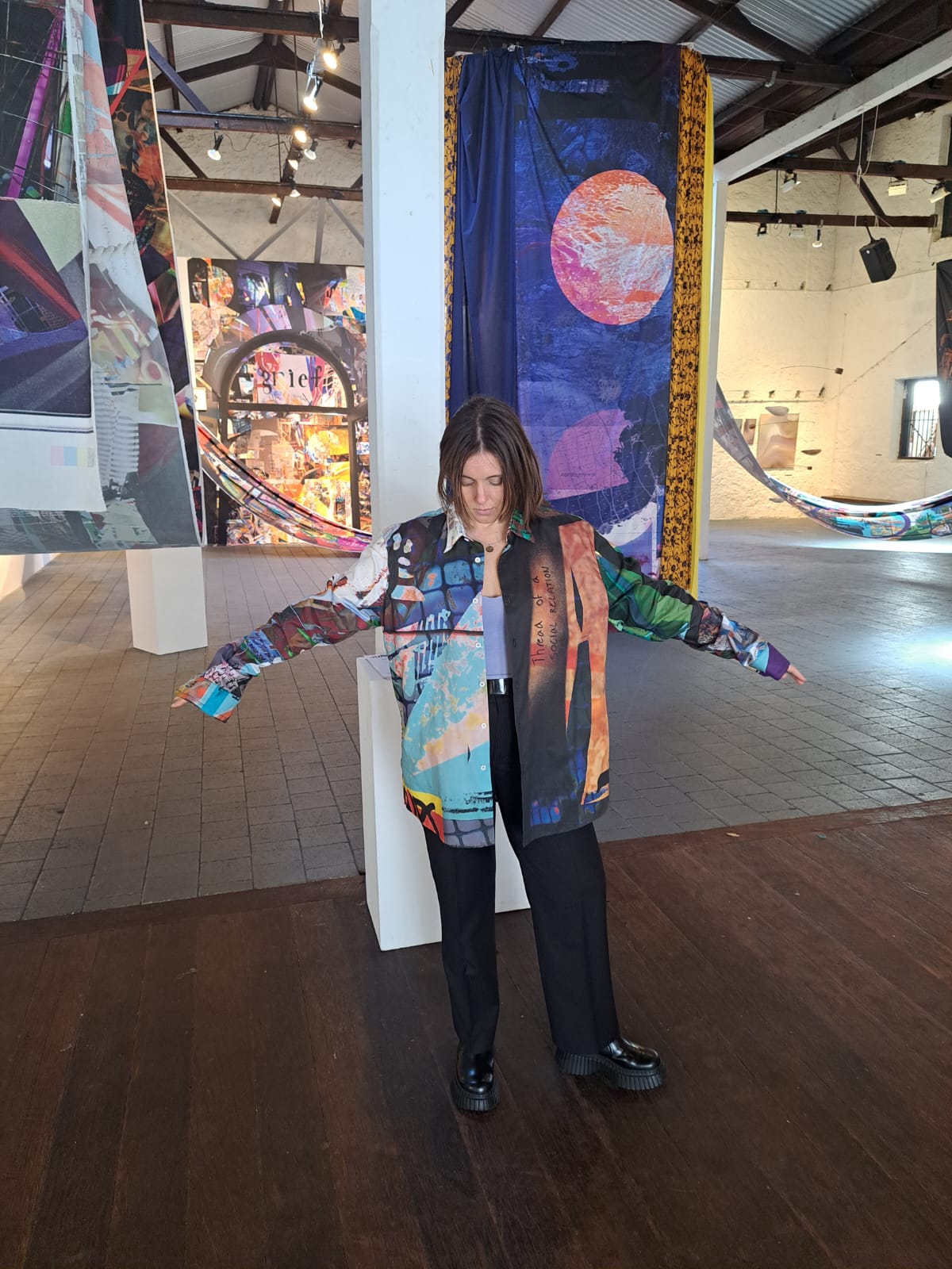 Person wearing a colorful longsleeved shirt in an art gallery with large tapestries hanging in the space.